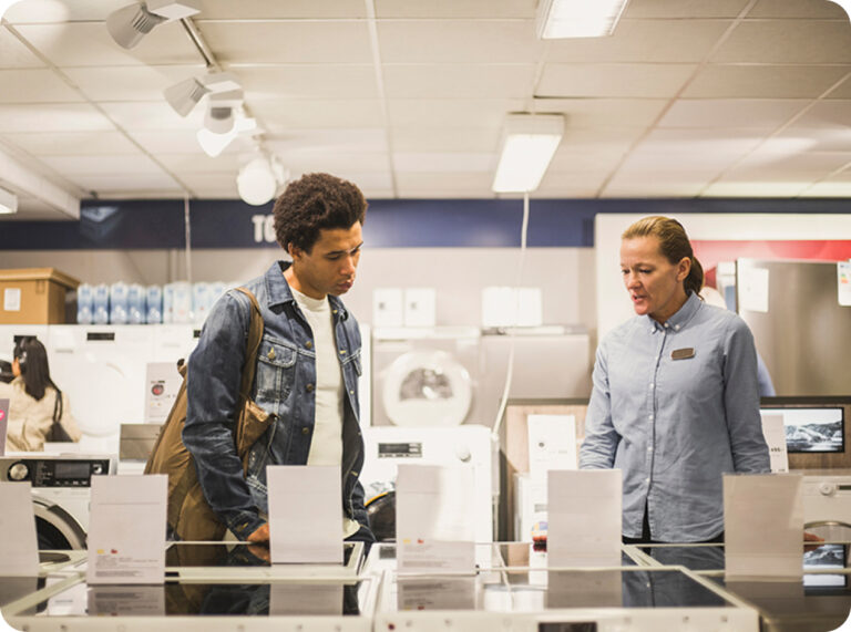 Två personer i en ljus butik, en man i jeansjacka granskar vitvaror och en kvinnlig säljare i ljusblå skjorta står bredvid