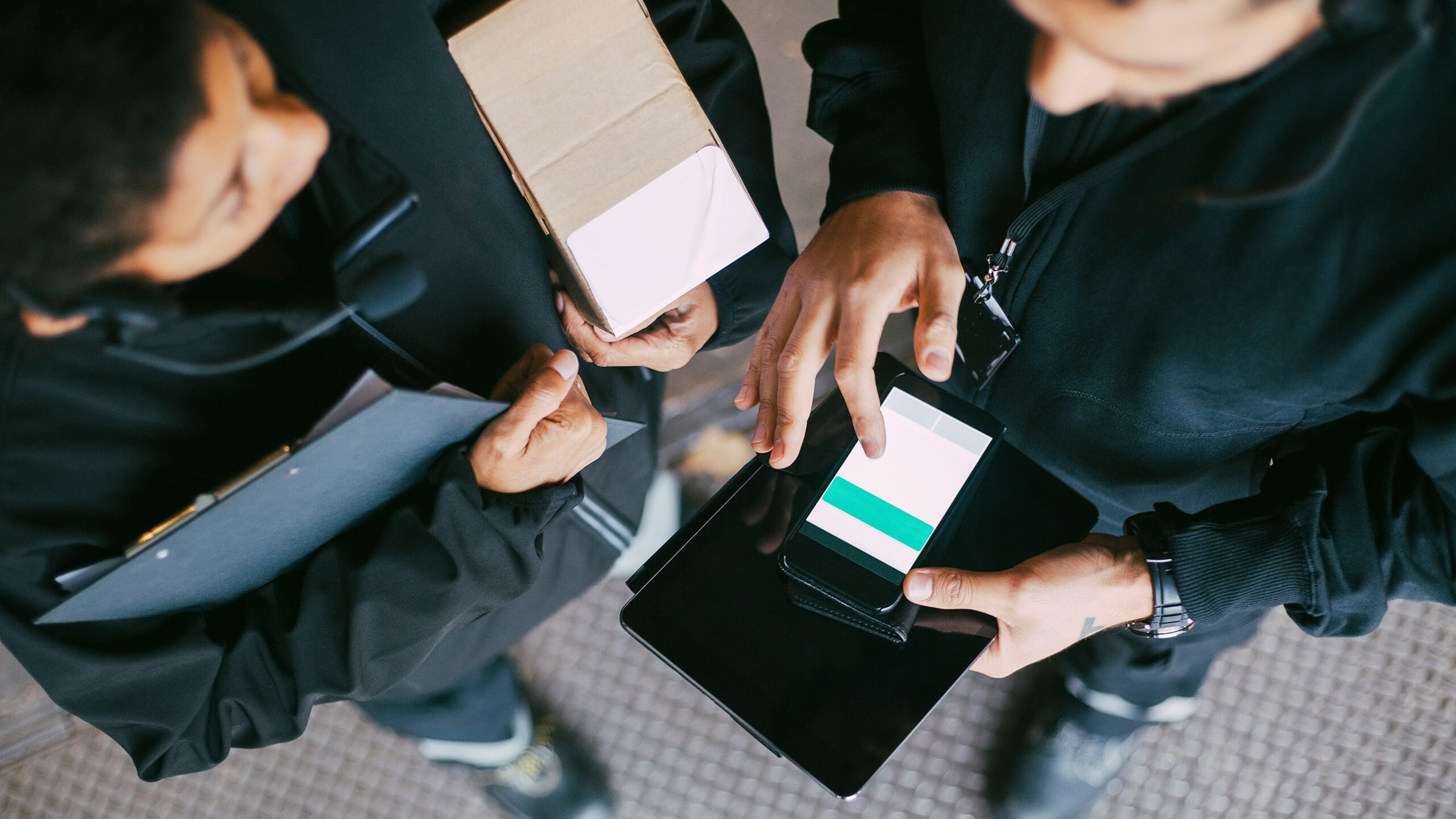 Ovanifrånbild på två personer i mörka arbetskläder, en registrerar order i mobil kopplad till Cash-IT:s kassasystem, den andra bär ett brunt paket