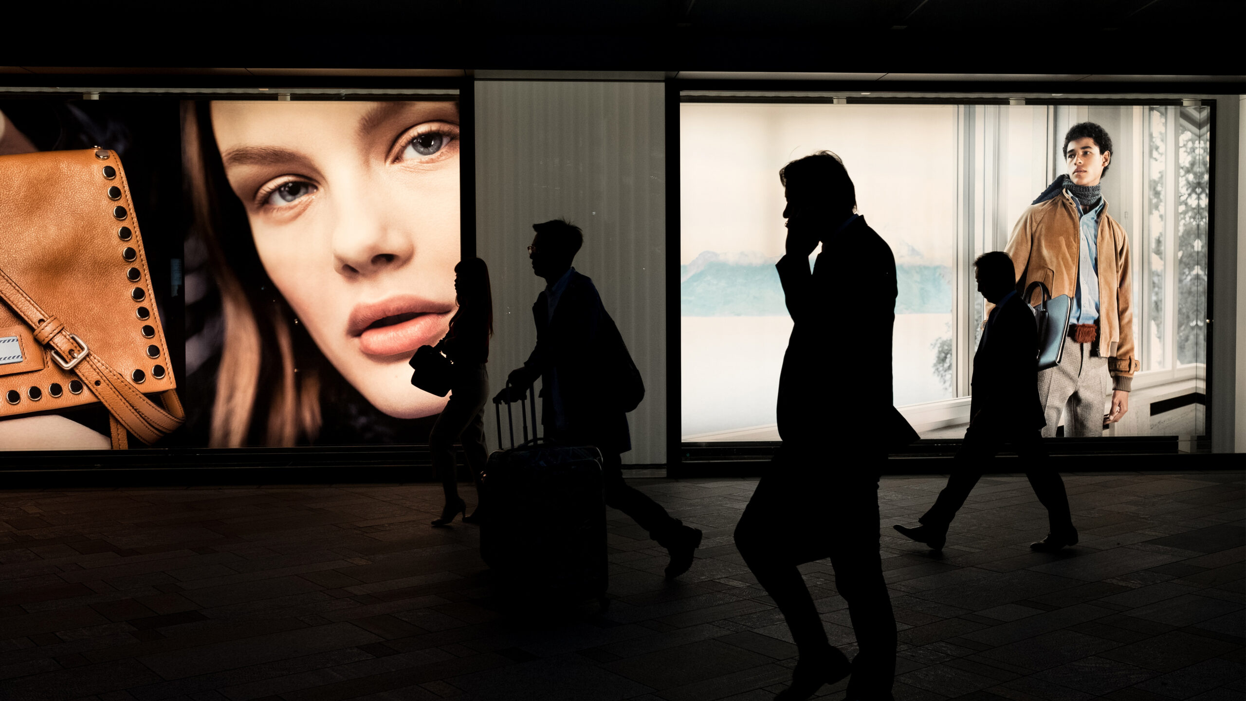 Silhuetter av forbipasserende mennesker foran opplyste reklameskilt med nærbilde på kvinne, brun veske og mann i moteriktig antrekk