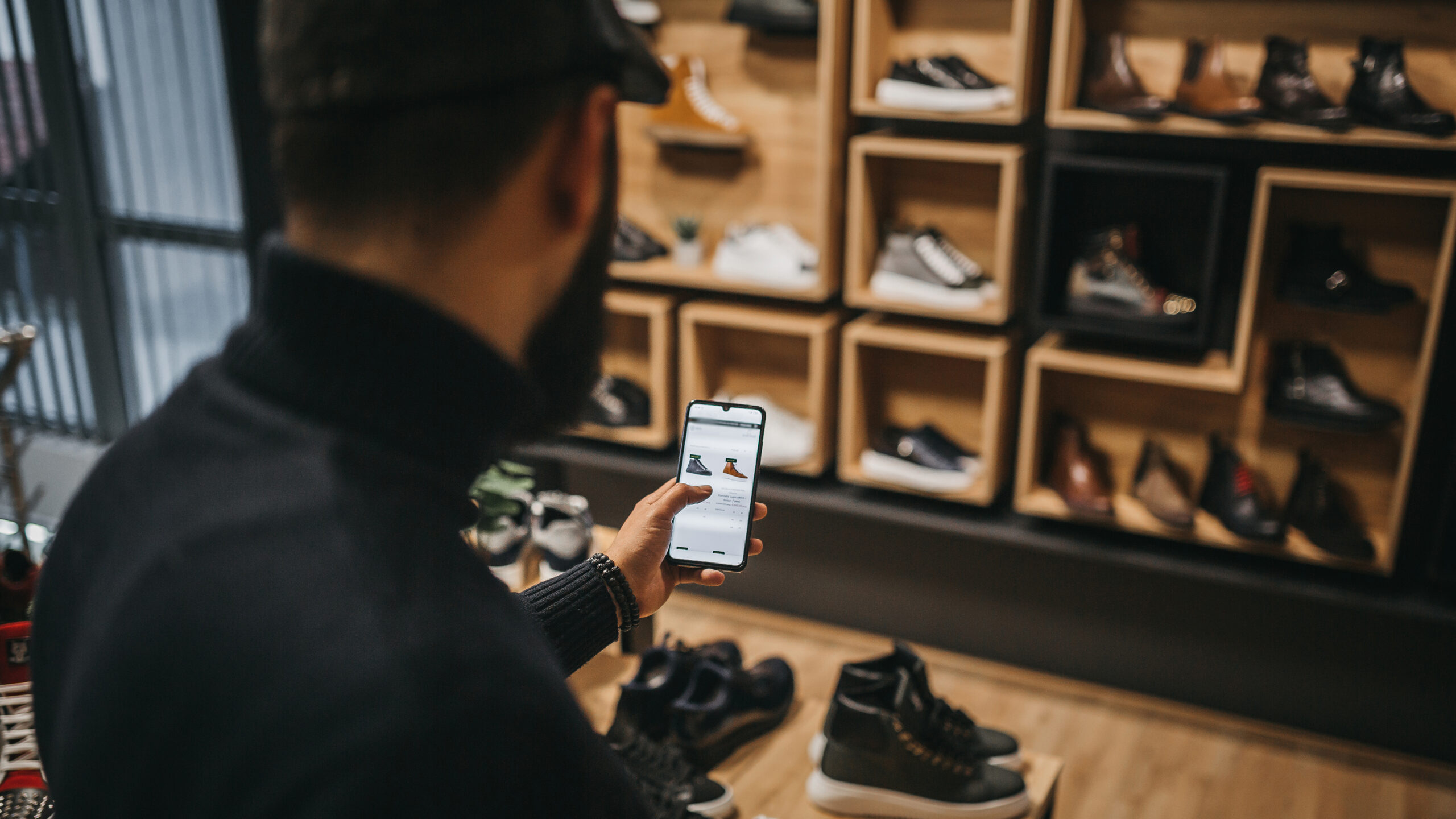 Digital betalingsløsning i mobilen brukes av person i mørke klær i en skobutikk med treinnredning og hyller fulle av sko