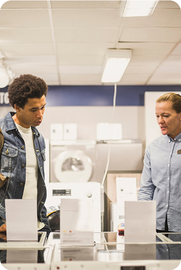 To personer i en velbelyst moderne butikk, en mannlig kunde i blå jeansjakke gransker hvitevarer mens en kvinnelig selger i lyseblå skjorte står ved siden av