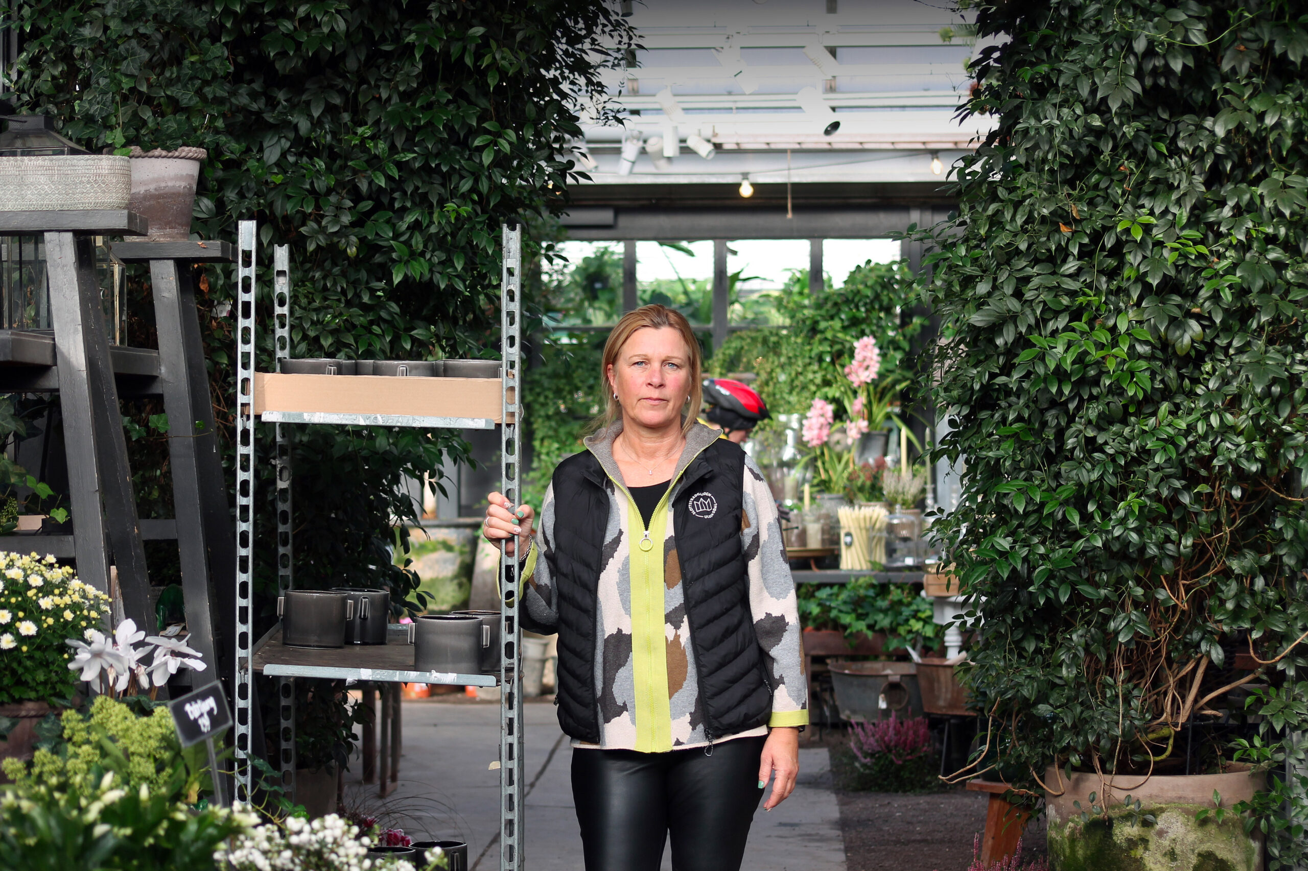Medelålders kvinna i mönstrad tröja och svart väst står i ett grönt växthus, omgiven av blommor, krukor och frodiga växter