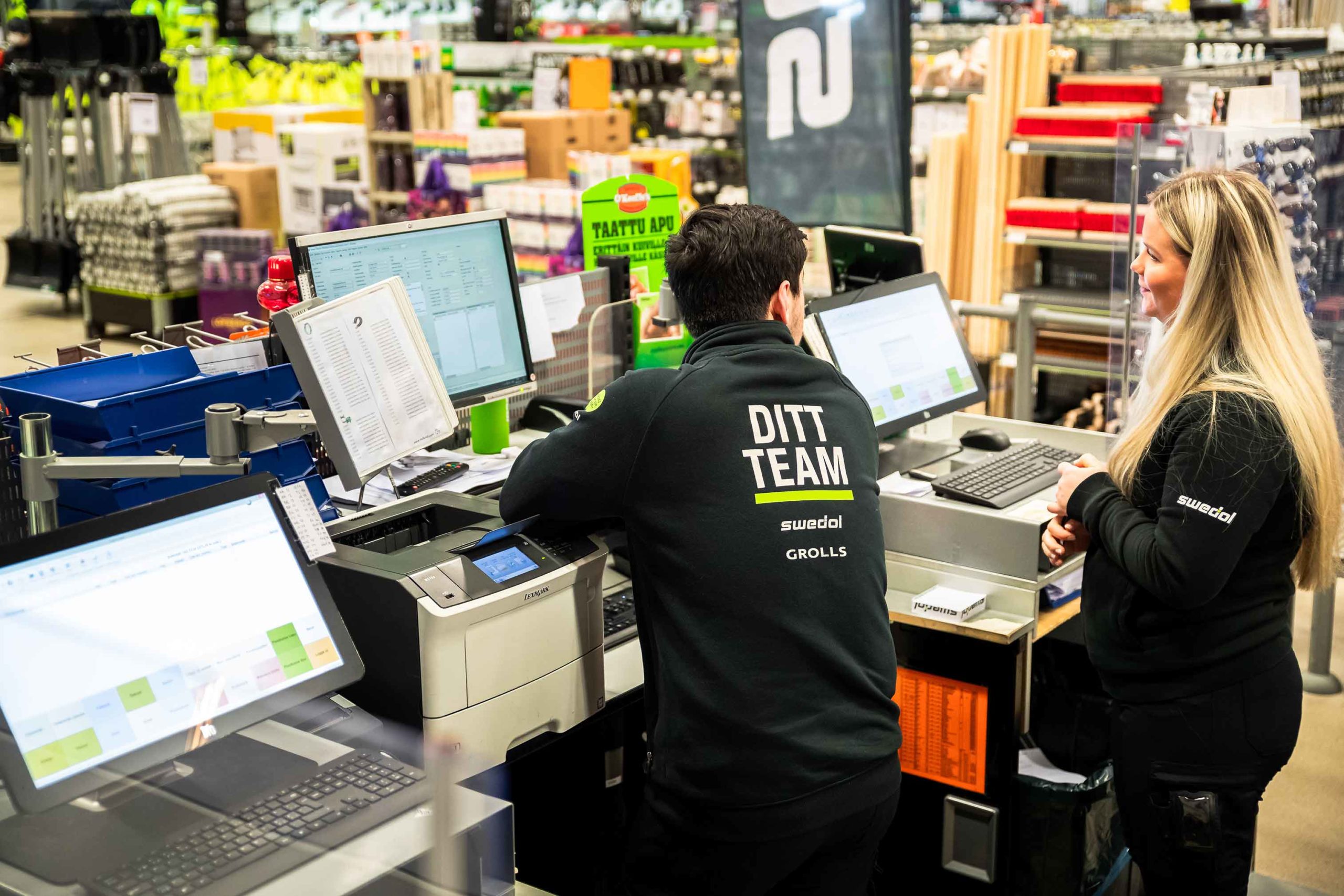 I en välbelyst butiksmiljö står två medarbetare i svarta Swedol-jackor vid kassadisk med flera skärmar, skrivare och pärmar