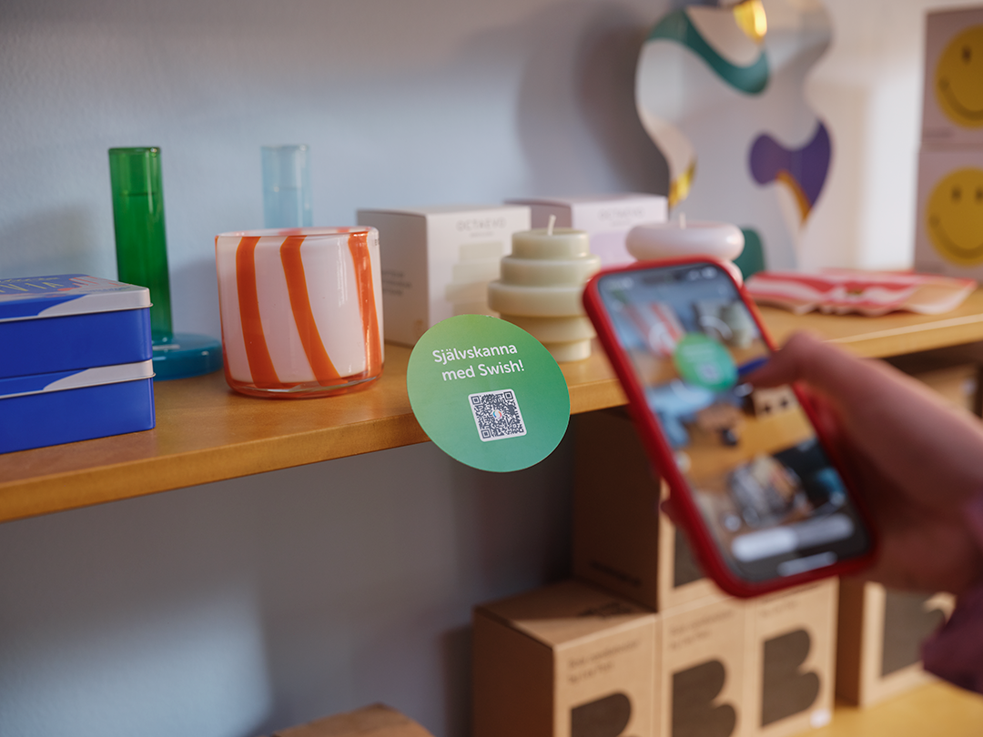 Kund skannar QR-kod för mobilbetalning med Swish på butikshylla med färgglada vaser och ljuslyktor