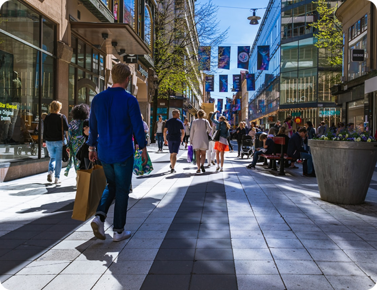 Människor promenerar längs en solbelyst shoppinggata med butiker, glasfasader och blomkrukor. I fokus syns en man i blå skjorta med en shoppingpåse