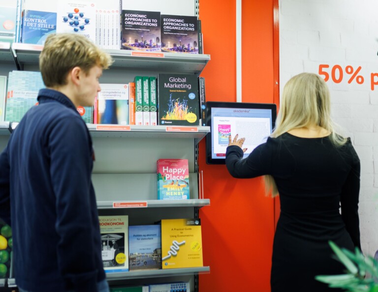 En ung kvinne i svart kjole interagerer med en veggmontert digital skjerm, ved siden av til venstre er det en bokhylle med bøker og en mann i mørkeblå genser observerer