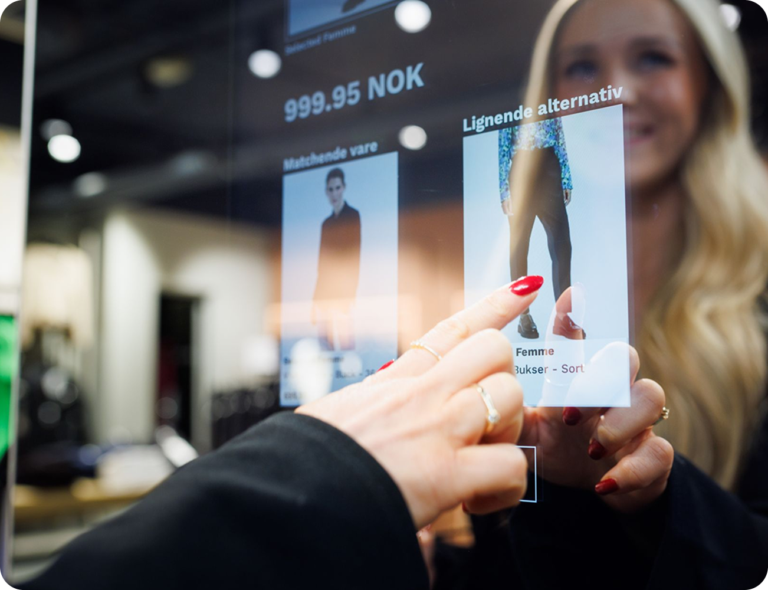 Interaktiv butikkskjerm med priser og produktvalg, en hånd med røde negler peker på et bilde av et par bukser