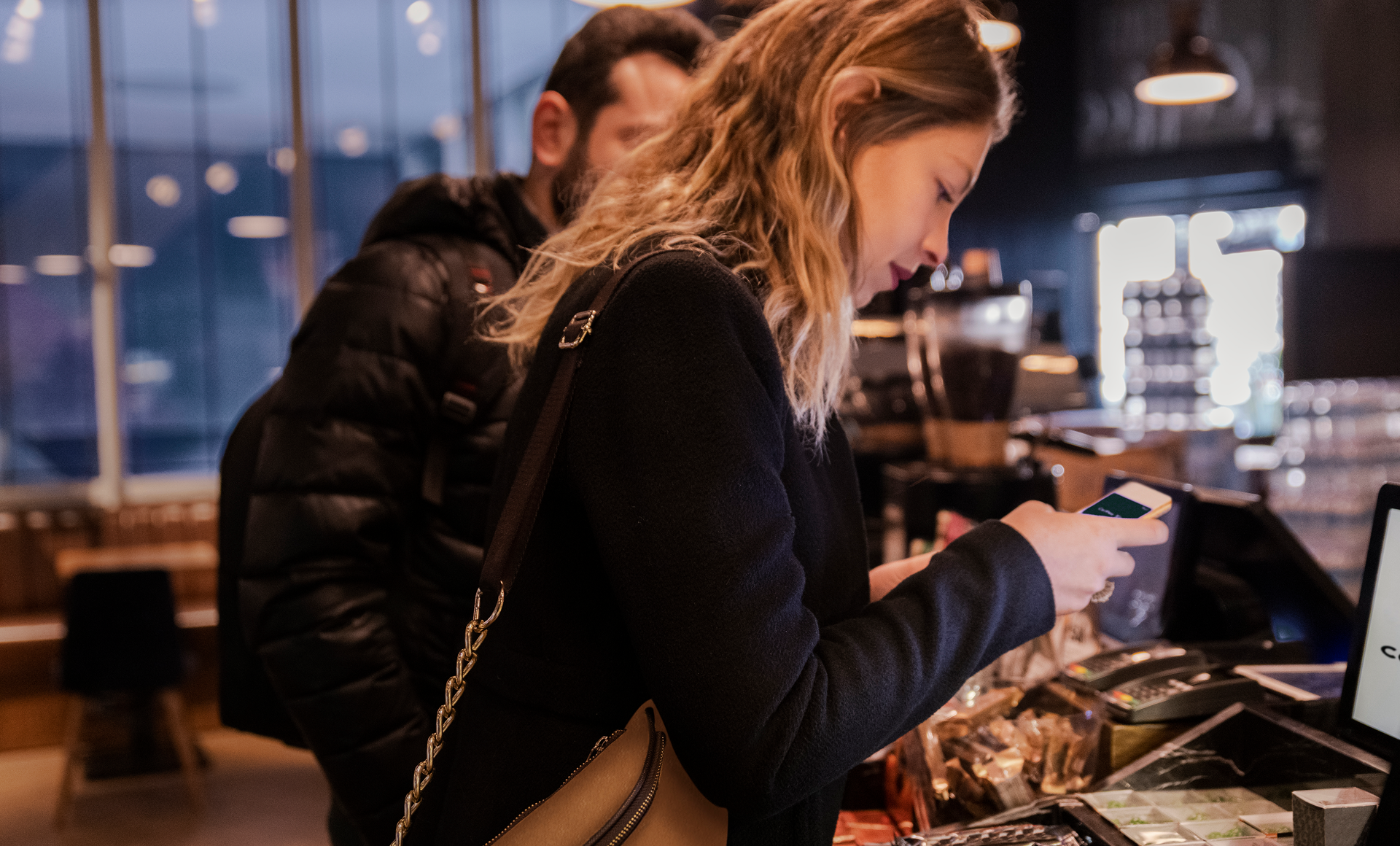 Ung kvinne holder frem mobil for digital betaling i kassen på kafé eller butikk, med en ventende kunde bak i dunkelt belyst butikk med mørk innredning