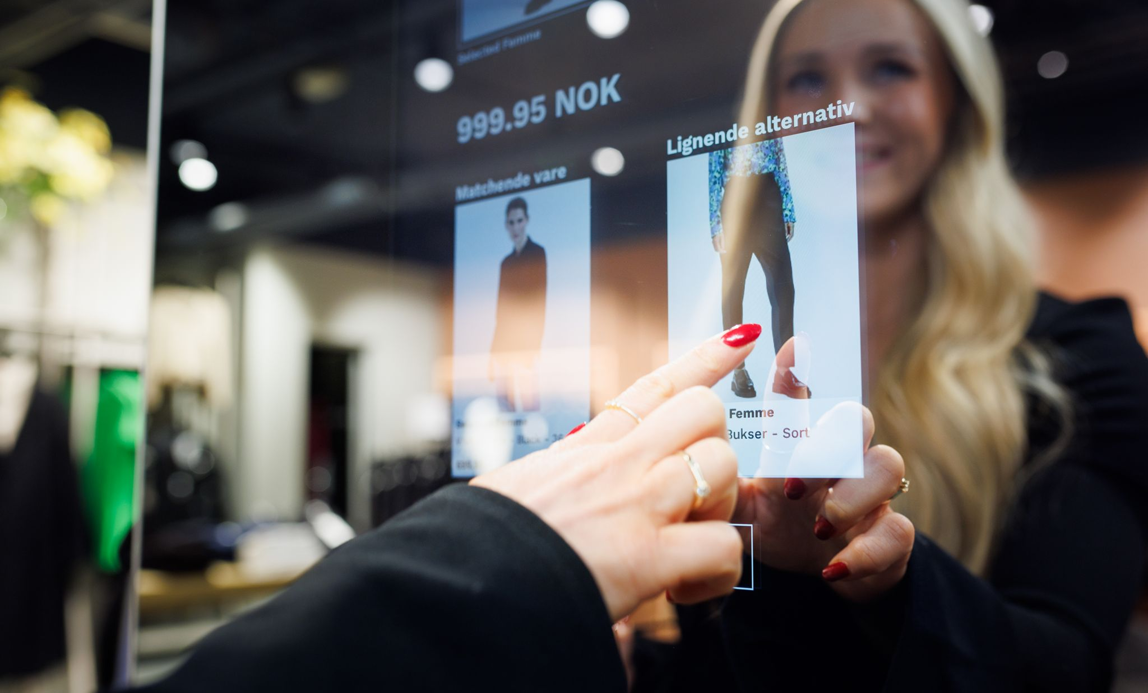 Ung blond kvinne foran en moderne interaktiv skjerm, der en hånd med røde negler peker på et produktbilde med et bilde av et par bukser